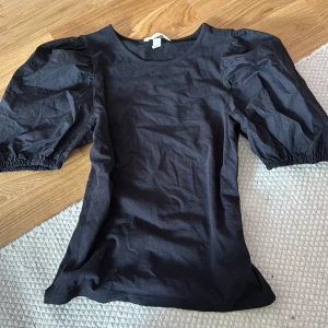 Svart topp med puffärmar - Snygg svart topp med puffiga kortärmar och rund halsringning. Toppens material känns mjukt och lätt, perfekt för att lyfta en enkel outfit. Ärmarna har resår vid avslutet för extra volym och en trendig siluett.