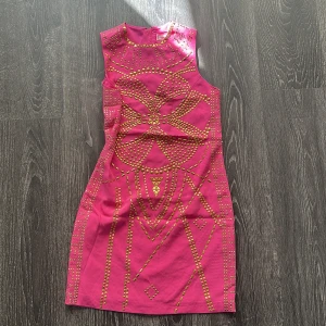 Rosa kort klänning med guldnitar - Snygg rosa kort klänning med coola guldnitar i geometriskt mönster framtill och längs sidorna. Klänningen är ärmlös och har en rak passform med rund hals. Dragkedja i guld hela vägen bak för en edgy vibe. Perfekt för dig som vill sticka ut med färg och detaljer.