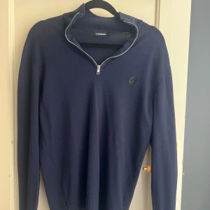 Marinblå half zip tröja J.Lindeberg - Mörkblå stickad tröja från J.Lindeberg med half zip och hög krage. Tröjan har diskret logga på bröstet och ribbade muddar vid ärmslut och nederkant. Perfekt för lager-på-lager och snygg till jeans eller chinos.