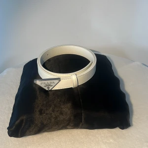 Vitt skärp från Prada i skinn - Snyggt vitt bälte från Prada i äkta skinn med en ikonisk trekantig silverfärgad logga framtill. Bältet har stilrena sömmar och en clean look som lyfter vilken outfit som helst. Perfekt accessoar för dig som gillar exklusiva detaljer och modern design.