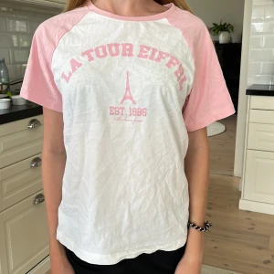Vit och rosa t-shirt La Tour Eiffel - Snygg t-shirt med vit bas och ljusrosa ärmar samt tryck framtill med texten 'La Tour Eiffel' och Eiffeltornet. Klassisk rund halsringning och avslappnad passform. Perfekt för dig som gillar Paris-vibes och mjuka färger.