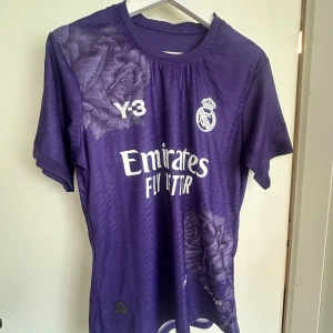 Lila Real Madrid Y-3 x Adidas Autentisk Kvalité - Säljer en limited edition Real Madrid fotbollströja i lila från Y-3 x Adidas. Tröjan har korta ärmar, blommigt mönster med rosor, och tryckta loggor från Real Madrid, Y-3 och Emirates. Materialet är lätt och andas, perfekt för match eller träning.