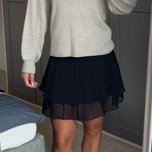 Svart volangkjol med prickigt mesh - Supersöt svart kortkjol med dubbla volanger och transparent mesh med diskret prickigt mönster. Kjolen har en flowy passform och resår i midjan för extra komfort. Perfekt att styla med en stickad tröja eller crop top.