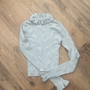 Ljusgrå ribbad polotröja med volang - Superfin ljusgrå polotröja med ribbad struktur och volangdetaljer vid krage och ärmslut. Tröjan har långärmade smala ärmar och en hög, rynkad polokrage som ger en extra stylish touch. Perfekt för dig som gillar en clean men ändå söt look.