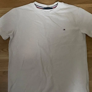 Vit t-shirt Tommy Hilfiger S - Vit t-shirt från Tommy Hilfiger i storlek S. Klassisk design med diskret logga på bröstet och rund hals. Materialet är bomull med en lätt textur som ger en cool känsla. Perfekt för dig som gillar stilrena och enkla plagg med en touch av premium. Storlek S passar 13-15 