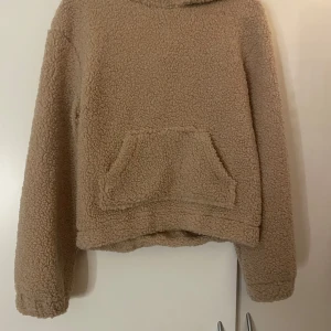Beige teddy hoodie från Hains - Supermysig beige teddy hoodie från Hains i storlek XS. Tröjan har huva, lång ärm och en stor magficka framtill. Perfekt för kalla dagar när du vill vara både chill och varm. Materialet är fluffigt och fleece-liknande, vilket gör den extra skön att ha på sig.