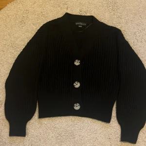 Svart ribbad kofta med stora knappar - Säljer en svart ribbad kofta med tre stora silvriga knappar framtill. Koftan har långärmade ballongärmar och är croppad i modellen. Materialet är stickat och känns mjukt och skönt mot huden. Perfekt för lager-på-lager och snygg till jeans.