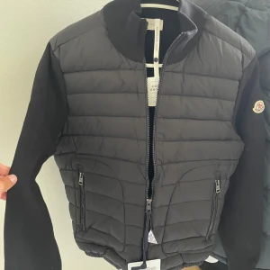 Moncler Cardigan - Snygg svart dunväst från Moncler med quiltad framsida och dragkedja. Västen har ribbad krage och två fickor med dragkedja. Klassisk Moncler-logga på ärmen och stilren design som passar till många outfits.