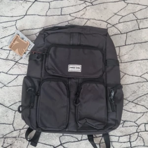 Svart ryggsäck från PRO-DG med USB-port - Snygg svart ryggsäck från PRO-DG med flera framfickor, laptopfack och USB-port för laddning. Tillverkad i slitstark polyester med vadderade axelband och rygg för extra komfort. Perfekt för skola eller cityhäng, med smarta detaljer och modern look.