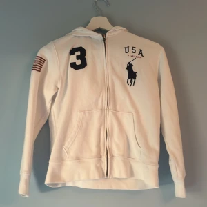 Vit hoodie från Polo Ralph Lauren - Vit hoodie från Polo Ralph Lauren med USA-tryck och stor svart polospelare på bröstet. Snygg amerikansk flagga på ena ärmen och siffran 3 i svart. Dragkedja framtill, huva och två fickor. Perfekt för dig som gillar sportig och klassisk stil. Bra skick men lite skrapad text och lite färg