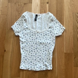 Vit blommig topp från H&M Divided - Supersöt vit topp med små blå blommor från H&M Divided. Modellen är ribbad med korta ärmar, v-ringning med spetskant och en liten rosett framtill. Perfekt för en söt och romantisk look. Materialet är mjukt och stretchigt, vilket gör den riktigt bekväm.