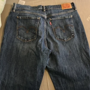 Levi's 501 blå jeans straight fit - Klassiska Levi's 501 jeans i blå denim med raka ben och normal passform. Jeansen har fem fickor, synliga nitar och den ikoniska läderpatchen bak i midjan. Snyggt slitna detaljer och orange sömmar ger en tidlös look.