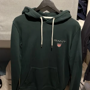 Mörkgrön hoodie från GANT med snörning - Säljer en mörkgrön hoodie från GANT i storlek M. Tröjan har vit snörning i huvan, broderad GANT-logga och sköld på bröstet samt en klassisk känguruficka framtill. Mjuk bomullskänsla och ribbade muddar vid ärmslut och nederkant. Perfekt för chill dagar.
