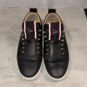 Christian Louboutin F.A.V. Spikade Slip-On Sneakers – Svart – Storlek 44 - Säljer ett par sneakers från Christian Louboutin, modell F.A.V. (Fique A Vontade), i storlek FR 44. Skorna är tillverkade i svart läder och har silverspikar istället för  skosnören.  Detaljer: – Storlek: FR 44 (EU 44 / US 11) – Svart läderöverdel – Silverfärgade spikdetaljer – Slip-on-design – Röd gummisula – Louboutin-logotyp på tunga och innerfoder  Skorna är i gott skick med spår av användning, främst på sulan. Ovandelen är välbevarad och utan större skador. Originalkartong medföljer ej.