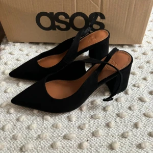 Svarta pumps med klack från ASOS - Säljer ett par eleganta svarta pumps från ASOS med spetsig tå och bred klack. Skorna har en stilren design med öppen häl och rem över foten. Perfekta för dig som vill ha en klassisk och trendig look.