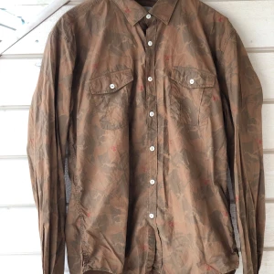 Barbour Beacon brun skjorta, storlek large - Brun, långärmad skjorta med ett unikt kamouflagemönster som inkluderar Barbour Beacon fyr. Skjortan har två bröstfickor med knappar och en klassisk krage. Knapparna är vita och skjortan har en slim fit passform i storlek Large. Använd en gång då den var för liten, tyvärr poppade en ärmknapp av p.g.a. det vilken jag inte hittade så den är ersatt med en lite mindre knapp som medföljde (sista bilden), i övrigt helt nyskick.