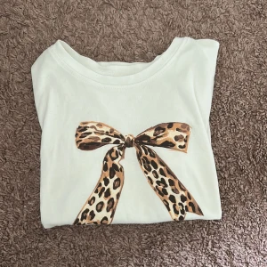 Vit t-shirt med leopardrosett - Vit t-shirt med korta ärmar och en stor rosett i leopardmönster framtill. Super skönt material och snygg till jeans💞