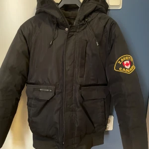 Svart vinterjacka med huva från Zavetti Canada - Svart vinterjacka från Zavetti Canada med huva och patch på ärmen. Jackan har dragkedja, snörning vid huvan och flera fickor framtill. Perfekt för kalla dagar.