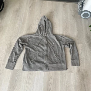 Ralph Lauren zip hoodie  - Säljer en grå hoodie från Polo Ralph Lauren med dragkedja och fickor framtill. Klassisk broderad logga på bröstet och justerbar huva. Perfekt för en avslappnad stil. Storlek M-L