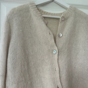 Beige/vit stickad kofta med knappar - Mysig beige stickad kofta med rund halsringning och stora knappar framtill. Koftan har långa ärmar och ribbade muddar vid ärmslut och nederkant. Perfekt att slänga på sig när det är kyligt ute över en klänning eller likande .