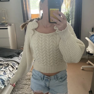 Vit stickad croppad tröja med knyt - Superfin vit stickad tröja med croppad passform och breda ärmar. Tröjan har ett snyggt flätmönster, volangdetaljer vid axlarna och knyt i ryggen som ger en unik look. Perfekt för dig som vill ha något gulligt men ändå trendigt.