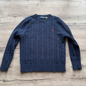 Stickad Polo Ralph Lauren tröja - Kabelstickad mörkblå tröja från Polo Ralph Lauren med rund halsringning och röd broderad logga på bröstet. Tröjan är långärmad och har ribbade muddar vid ärmslut och nederkant. Perfekt för dig som gillar klassisk och stilren look. Nästan helt ny endast använd 2 gånger