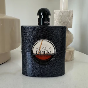 Yves Saint Laurent Black Opium Eau de Parfum 90ml - Säljer en ikonisk parfym från Yves Saint Laurent – Black Opium. Flaskan är svart med glittrig yta och rund transparent detalj i mitten där parfymens bärnstensfärgade vätska syns. Rymmer 90 ml och har en elegant svart kork.