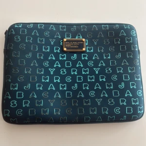Blå datorväska Marc by Marc Jacobs - Säljer en blå datorväska från Marc by Marc Jacobs med coolt bokstavsmönster i turkost över hela väskan. Väskan har en guldig logoplatta framtill och stängs med dragkedja. Perfekt för att skydda din laptop med stil. Storlek 34cm * 25cm