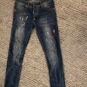 Dsquared2 skinny jeans blå, stl 30 - Snygga blå skinny jeans från Dsquared2 med slitna detaljer och coola färgstänk. Klassisk femficksmodell med läderpatch bak i midjan. Jeansen har en smal passform och är tillverkade i stretchig denim för extra komfort.
