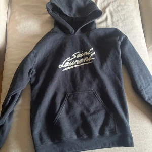 Svart Saint Laurent hoodie  - Svart hoodie från Saint Laurent med vit logga framtill. Klassisk känguruficka och huva, perfekt för en chill och stilren look. Mjuk bomullskänsla och långärmad modell som passar året runt.
