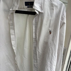 Vit skjorta från Ralph Lauren - Klassisk vit skjorta från Ralph Lauren med broderad logga på bröstet. Skjortan har lång ärm, knappar framtill och är i bomull. Slim fit-modell som passar perfekt till jeans eller chinos. Perfekt till sommaren. Medium slim fit 