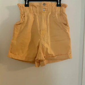Högmidjade shorts med volang - Snygga högmidjade shorts i apricot färg med volangkant upptill och uppvikta benslut. Shortsen har två fickor fram och stängs med knappar och dragkedja. Perfekta för varma sommardagar och ger en fräsch look till din outfit.