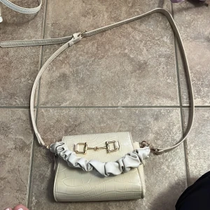 Beige axelväska med krokoprägling - Snygg beige axelväska i krokopräglat syntetmaterial med guldfärgade metalldetaljer och en scrunchie-inspirerad handrem. Väskan har justerbar axelrem och magnetlås. Perfekt storlek för mobil, plånbok och småsaker.