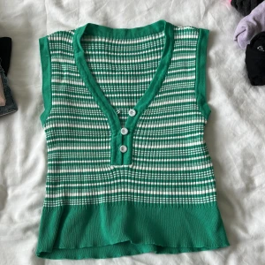 Grön och vit randig ribbad linnetopp - Säljer en grön och vit randig ribbad linnetopp med v-ringning och tre vita knappar framtill. Toppen är ärmlös och har en tight passform med stretchigt material. Perfekt för dig som gillar retrovibbar och vill sticka ut lite extra.