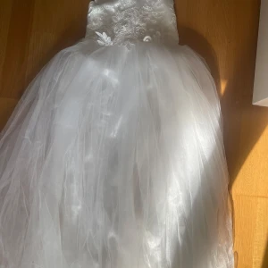 Vit långklänning med spets och rosett - Elegant vit långklänning med vacker spets upptill och broderade blommor. Kjolen är i flera lager av tunn tyll som ger en drömmig känsla. Baktill finns en stor rosett i satin. Perfekt för speciella tillfällen. Storlek 8-10år. Köpt i USA