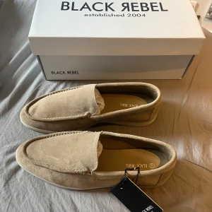 Beige mocka loafers från Black Rebel - Snygga beige loafers i mocka från Black Rebel. Skorna har en klassisk design med rund tå och platt sula, perfekta för en stilren look. Mjukt material och diskreta sömmar ger en clean känsla.