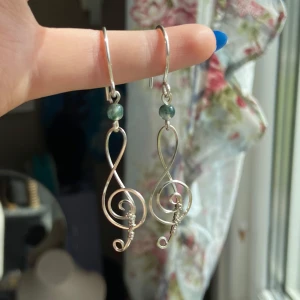 Silvriga örhängen med notdesign - Unika örhängen i form av g-klavar, handgjorda i silvrig metalltråd med små gröna pärlor upptill. Perfekta för dig som älskar musik och vill sticka ut med en kreativ och personlig stil. Lätta att bära och riktigt snygga till både vardagslooken och när du vill vara extra cool.