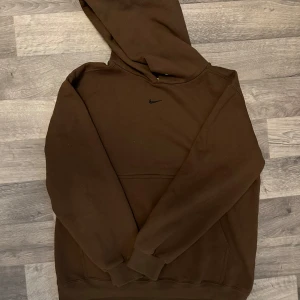 Brun hoodie från Nike med huva - Säljer en brun hoodie från Nike med klassisk svart swoosh-logga på bröstet. Tröjan har huva, magficka och långa ärmar med muddar. Perfekt för chill dagar eller när du vill ha en enkel och stilren look.