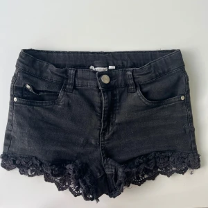 Svarta lågmidjade jeansshorts med spetskant - Snygga lågmidjade svarta jeansshorts med dekorativ spetskant längst ner. Shortsen har klassisk knapp och dragkedja framtill. De har en liten slitning på spetskanten se bild 2, men inget som märks eller syns. 
