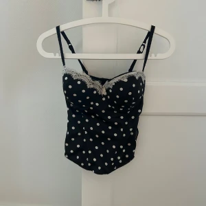  prickig korsettopp - Säljer en svart korsettopp från F&F med vita prickar och dekorativ vit spets längs bysten. Toppen har justerbara axelband och en figurnära passform. Ej vadderad i kuporna!! Säljer bara för annonsens pris 