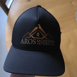 Svart keps Aros Moderna AB Flexfit - Svart keps med guldtryck från Aros Moderna Aktiebolag. Klassisk modell med böjd skärm och Flexfit 110-teknik för skön passform. Tillverkad i 98% polyester och 2% elastan. Snygg och stilren med logga framtill.