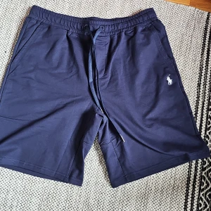 Blå shorts från Polo Ralph Lauren - Snygga marinblå shorts från Polo Ralph Lauren med klassisk broderad logga på benet. Mjukt bomullsmaterial, elastisk midja med snörning och två sidofickor. Perfekta för chill dagar eller när du vill ha en clean och stilren look. Stl M men passar även S