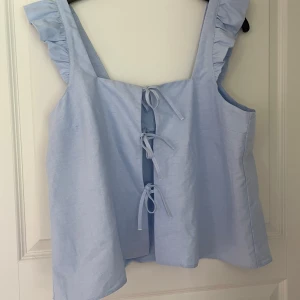 Ljusblå topp från Djerf Avenue - Supersöt ljusblå topp från Djerf Avenue med volangdetaljer på axelbanden och tre knytningar framtill. Toppen är ärmlös och har en luftig. Perfekt för varma dagar och ger en fräsch vibe till din outfit. 🫶🏼🫶🏼🫶🏼 aldrig använd.