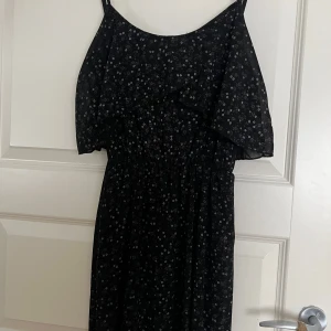 Svart blommig långklänning - Svart kortklänning med små vita blommor, offshoulder och volangdetalj upptill. Klänningen har smala axelband och ett luftigt, flowy tyg som ger en skön känsla. Perfekt för dig som gillar romantisk och stilren look.