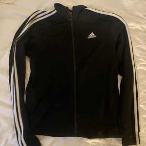 Svart träningsjacka med dragkedja från Adidas - Svart träningsjacka från Adidas med vita ränder längs ärmarna och dragkedja framtill. Klassisk Adidas-logga på bröstet. Jackan har lång ärm och en sportig passform, perfekt för träning eller vardagsstil. står ingen storlek men skulle gissa på M eller L?
