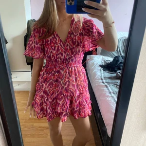 Zara klänning  - Säljer en färgstark kortklänning i rosa och rött med mönster, v-ringad hals och volangdetaljer. Klänningen har korta ärmar och smockad midja som ger en fin passform. Perfekt för sommarens tillfällen!