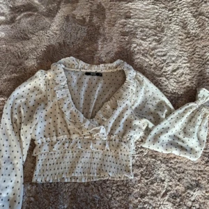 Vit blus med volang  - Supersöt vit blus från Bik Bok med svarta prickar, volangkant längs v-ringningen och ärmarna och smockad midja. Långa ärmar och lätt transparent material. Perfekt att styla med jeans eller kjol!