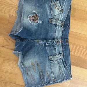 Blå jeansshorts med nitar och slitningar - Snygga blå lågmidjade jeansshorts från MET med råa kanter, slitningar och coola nitar framtill. Shortsen har detaljerade sömmar, dekorativa knappar och patch med leopardmönster baktill.  Storlek 34 i jeansstorlekar så skulle säga att de passar en M/L❤️‍🔥