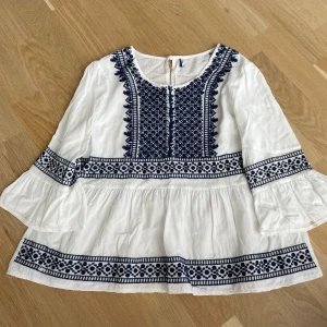 Vit blus med blå broderier - Supersöt vit blus med mörkblå broderade detaljer och mönster framtill, på ärmarna och nertill. Blusen har vida, långa ärmar och en lätt rynkad, flowy passform. Perfekt för dig som gillar boho-stil och snygga detaljer.