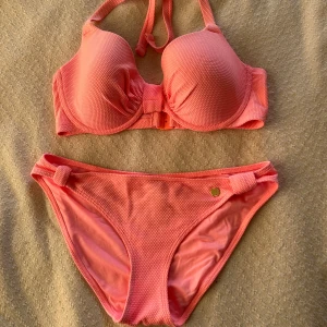 Rosa bikini med rosett och struktur - Säljer en snygg rosa bikini med vadderad bygel-bh och knyt i nacken. Bikinin har en liten rosett framtill och dekorativa knutar på trosorna. Materialet är strukturerat och känns lyxigt. Perfekt för stranden eller poolen!